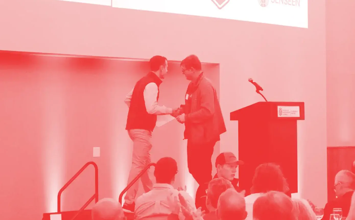 Two men standing on a stage in front of a crowd shake hands.